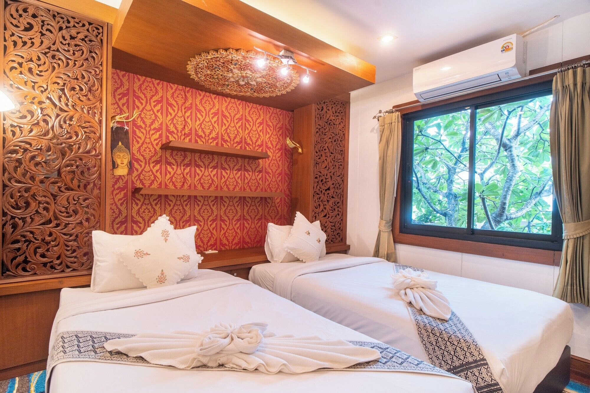 Een hotelkamer met twee bedden, gedetailleerd houten paneelwerk, een Boeddhabeeld en een raam met uitzicht op bomen.