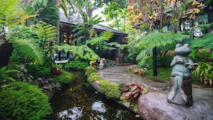 Un jardin luxuriant avec un sentier en pierre, un petit étang et un bâtiment traditionnel doté d’un toit en tuiles.