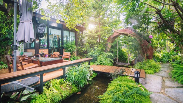 Un espace extérieur aménagé avec des tables et des chaises en bois, entouré d’une végétation luxuriante et agrémenté d’un petit élément décoratif aquatique.