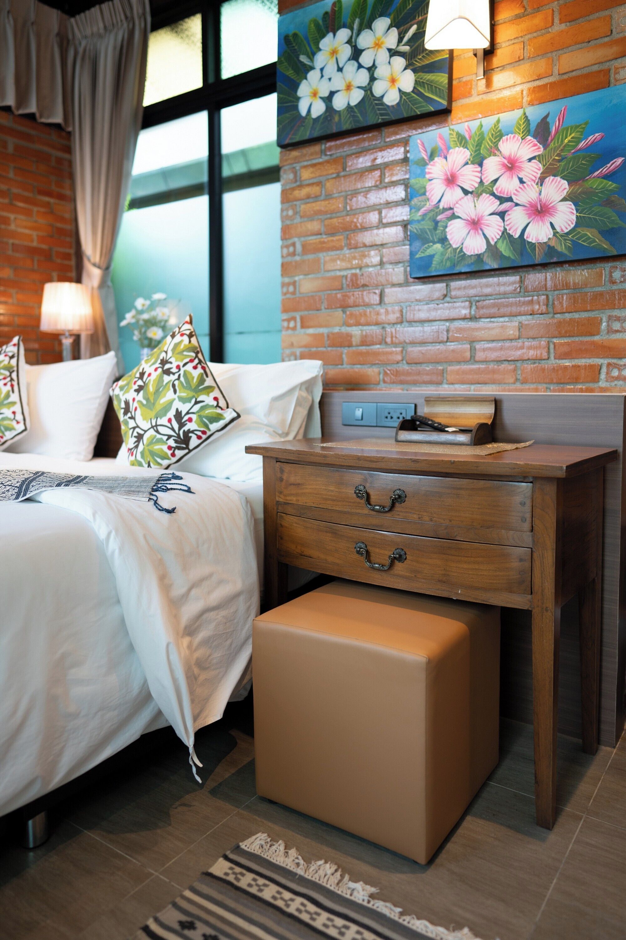 Un dormitorio con pared de ladrillo, cama con ropa de cama blanca, mesita de noche de madera y un pequeño cofre de madera.