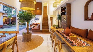Amplio espacio interior con una mesa de madera, sillas y un largo banco. Se ve una escalera que lleva a un nivel superior, y una planta grande en el centro.