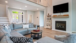 A spacious living room with a large sectional sofa, a fireplace, a flat-screen TV, and a staircase leading to an upper level. - NEW Build Keuka Lakefront | Sleeps 16 | Dock/Kayak (Penn Yan)