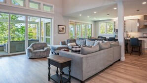 A spacious living room with a large sectional sofa, a small round table, and a view of the outdoors through large windows. - NEW Build Keuka Lakefront | Sleeps 16 | Dock/Kayak (Penn Yan)