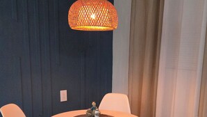 A dining area with a round table, four chairs, a hanging light fixture, and a wall with a dark blue panel and a curtain.