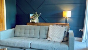 Un salon moderne avec un canapé gris clair, une table d’appoint en bois surmontée d’une lampe et un vase décoratif.