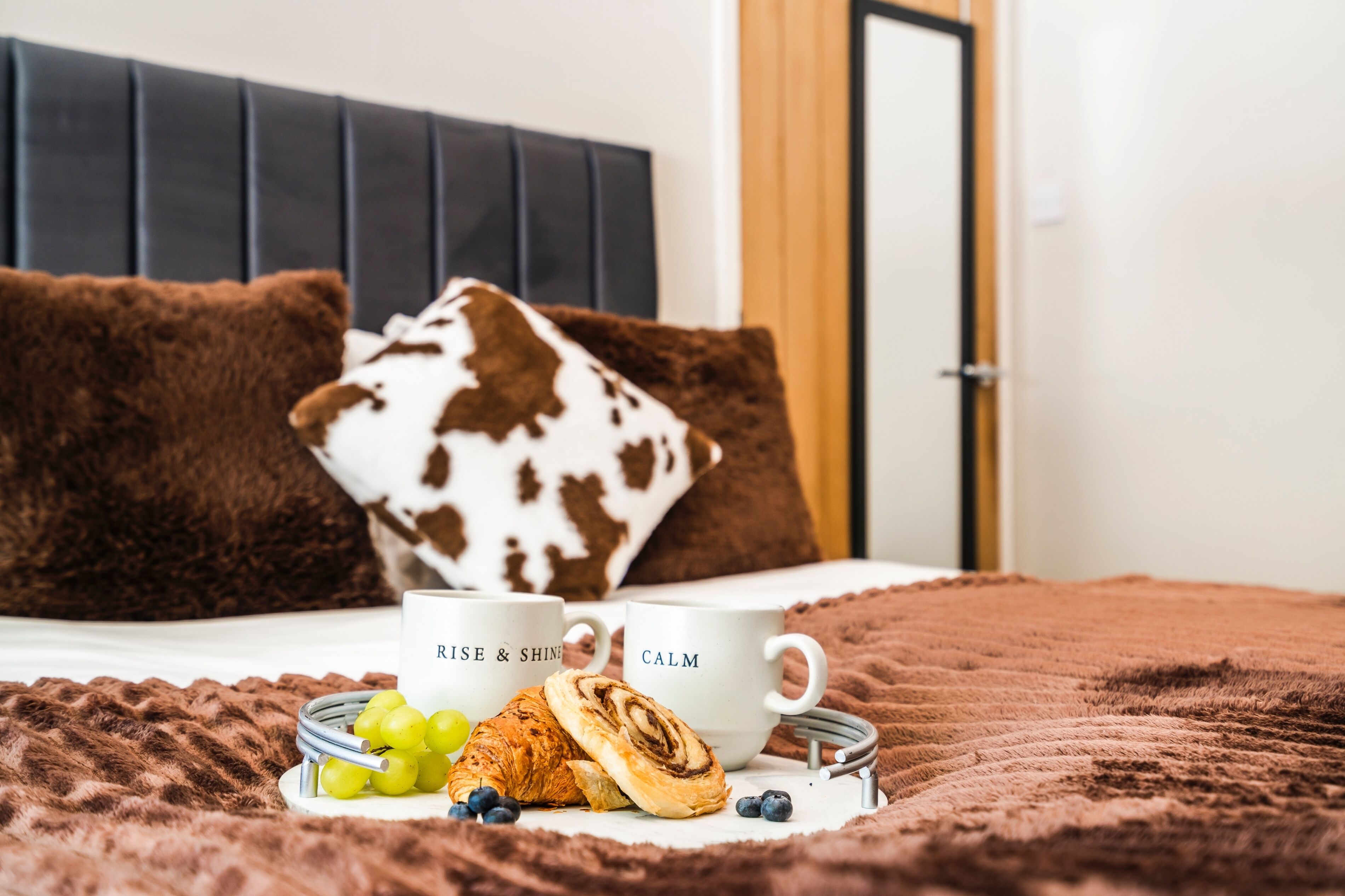 Ein Bett mit einer braunen Decke, zwei weiße Tassen mit Text, ein Teller mit Trauben und Gebäck sowie ein braun-weißes Kopfkissen.