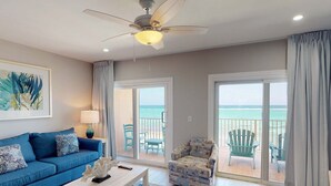 Un salon avec un canapé bleu, un fauteuil à motifs, une table basse et un ventilateur de plafond. Il y a un balcon avec vue sur l’océan.