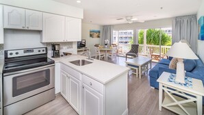 Une cuisine moderne avec des meubles blancs, un four encastré et un îlot central doté d’un évier. On y trouve également un coin repas avec une table et des chaises, un canapé bleu et un balcon offrant une vue sur la verdure.