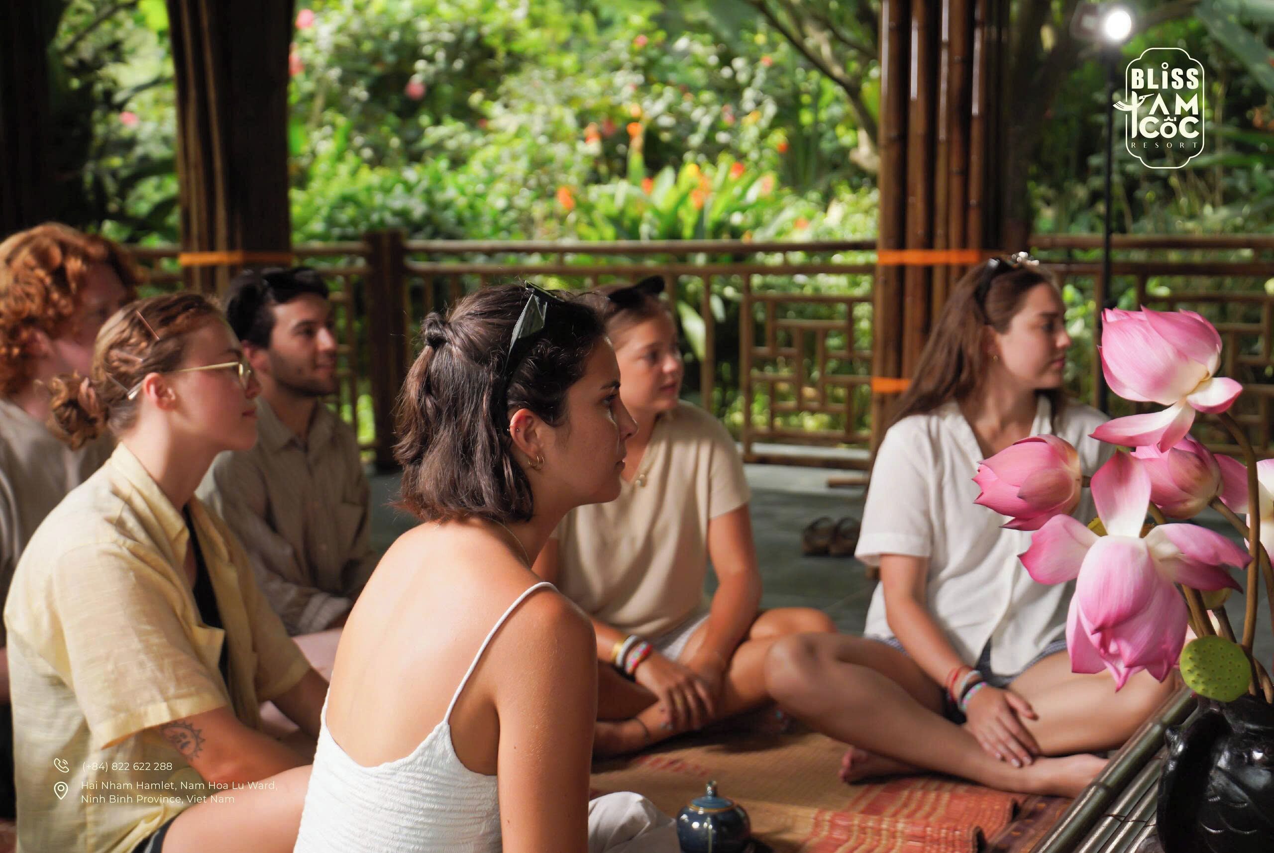 A group of people sitting on the floor in a covered area with bamboo structures and greenery in the background.