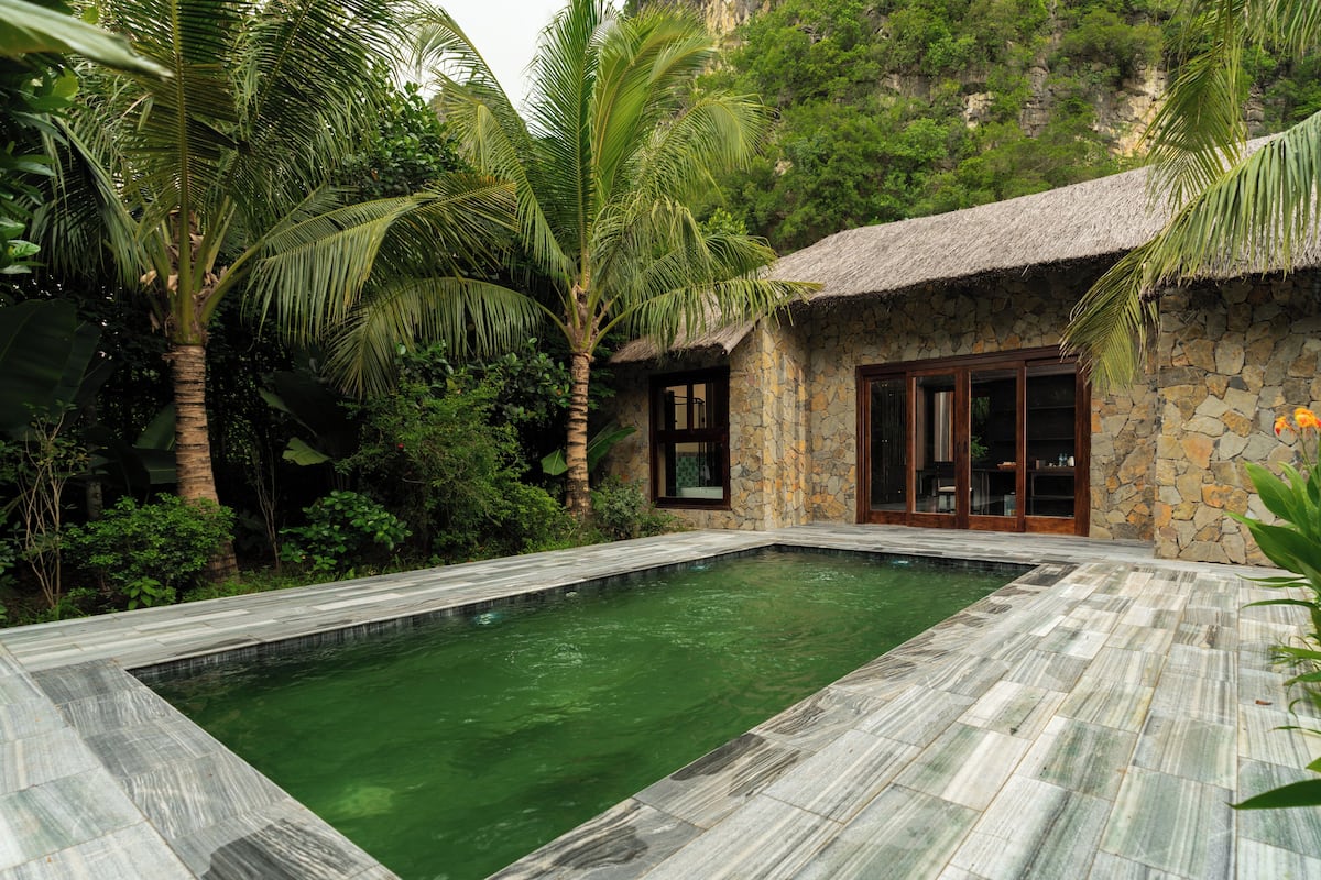Una casa de piedra con techo de paja, piscina y exuberante vegetación tropical.