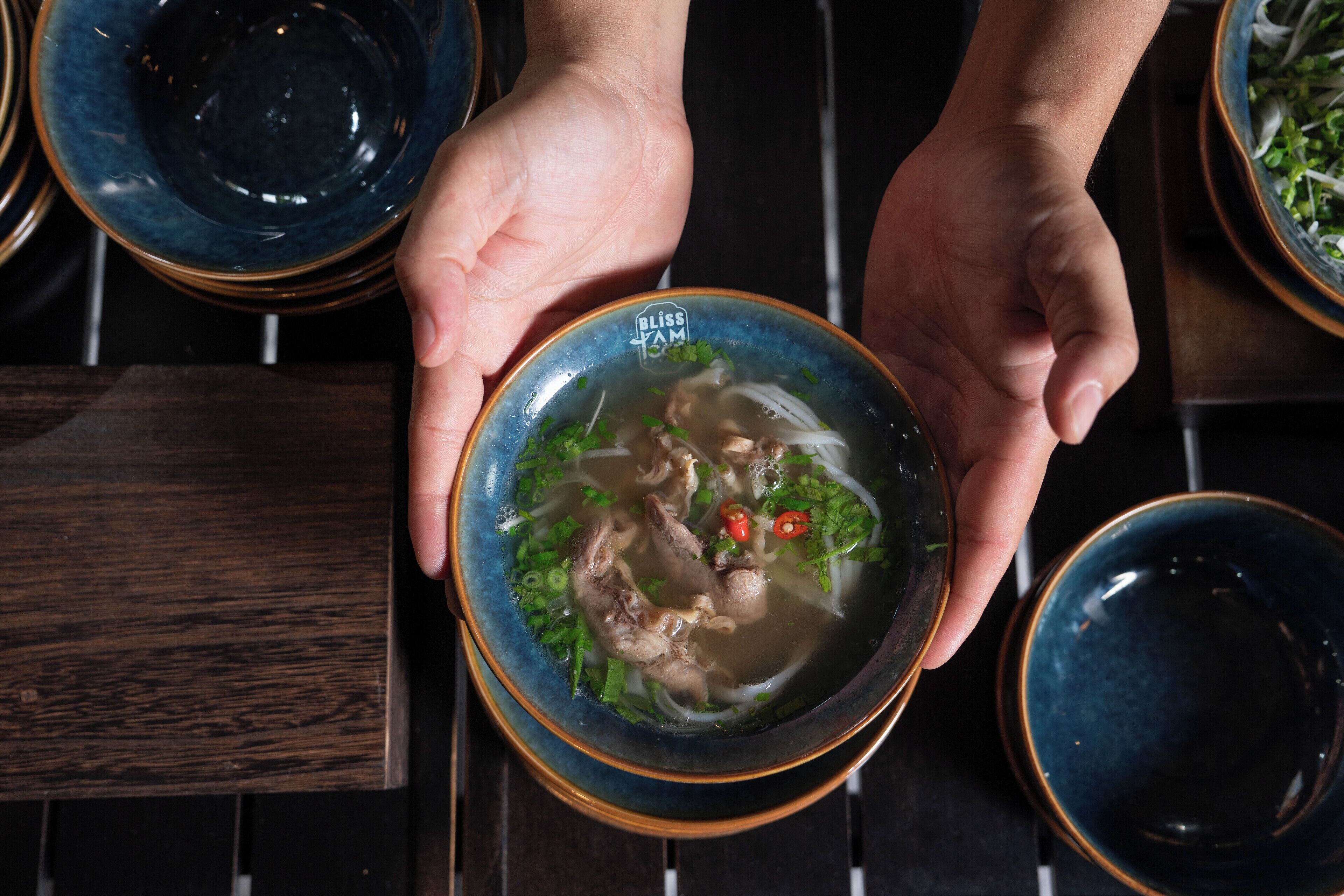 A person holding a bowl of soup with visible ingredients like noodles, herbs, and meat.