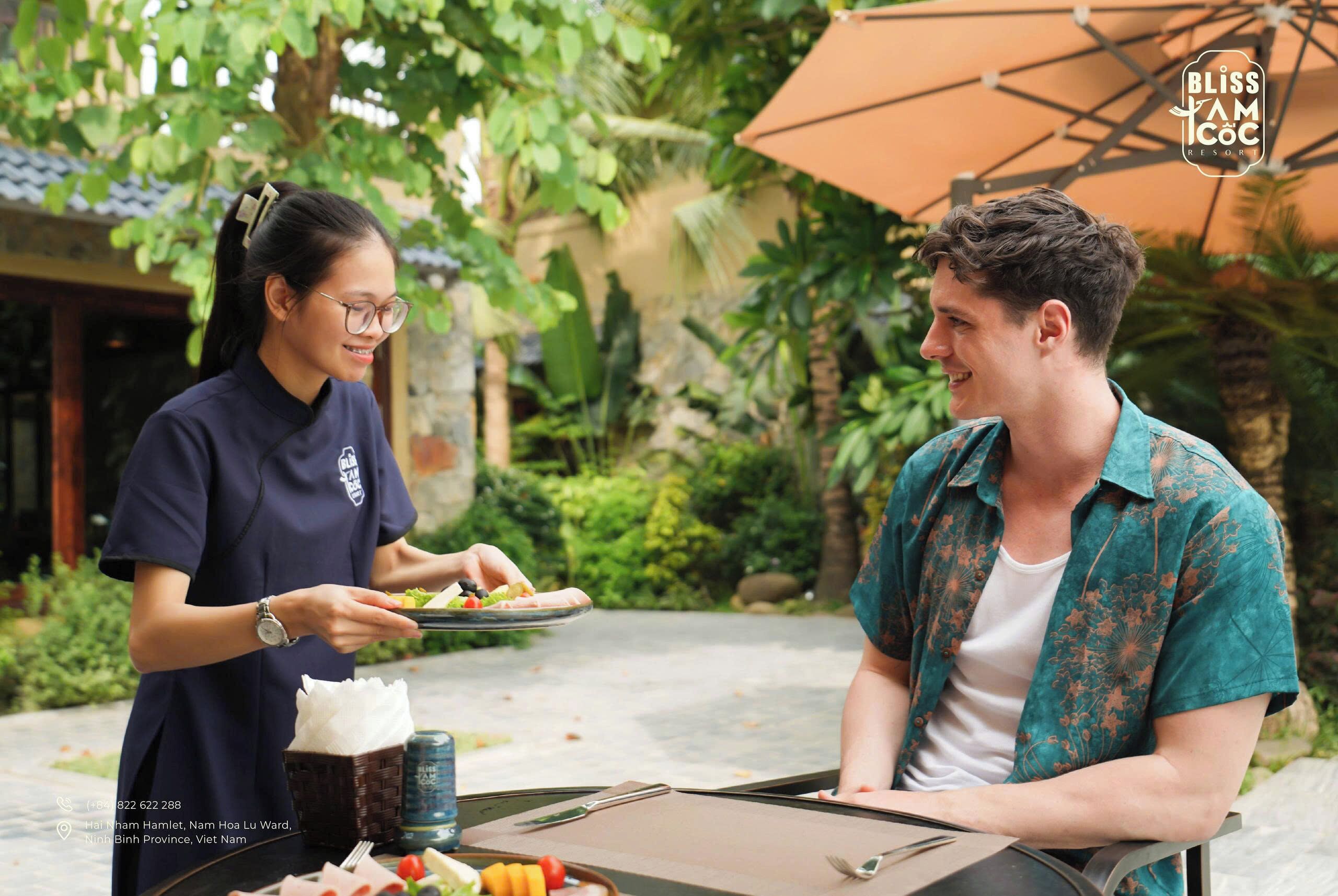 A person serving food to another at an outdoor table with a garden view, a table umbrella, and a menu.