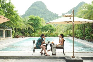 A poolside dining area with two people seated at a table, surrounded by lush greenery and a mountain in the background. - Bliss Tam Coc Resort Ninh Binh (Nam Hoa Lu)