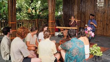 Eine Gruppe von Personen sitzt auf dem Boden in einer Bambuskonstruktion und lauscht einer Person, die ein traditionelles Musikinstrument spielt.