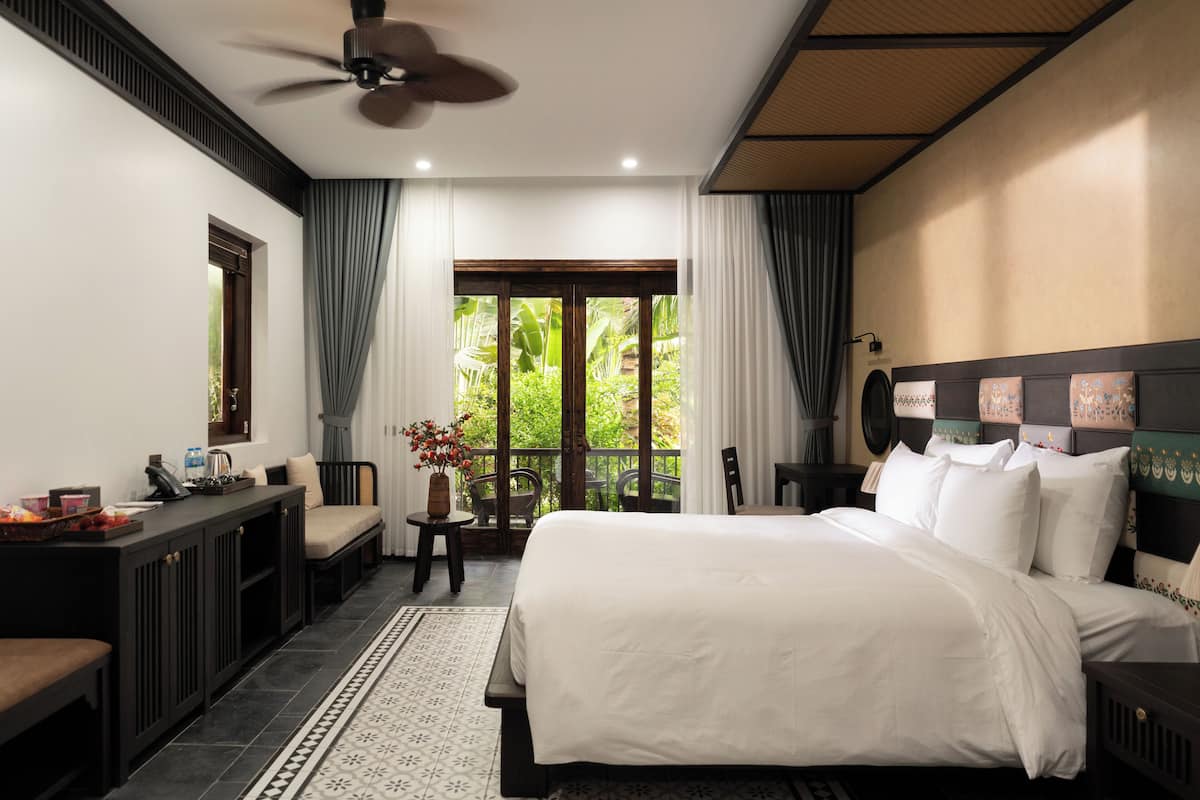 Un dormitorio con una cama grande, un escritorio y una silla. Hay una ventana con cortinas y vistas al verdor exterior.