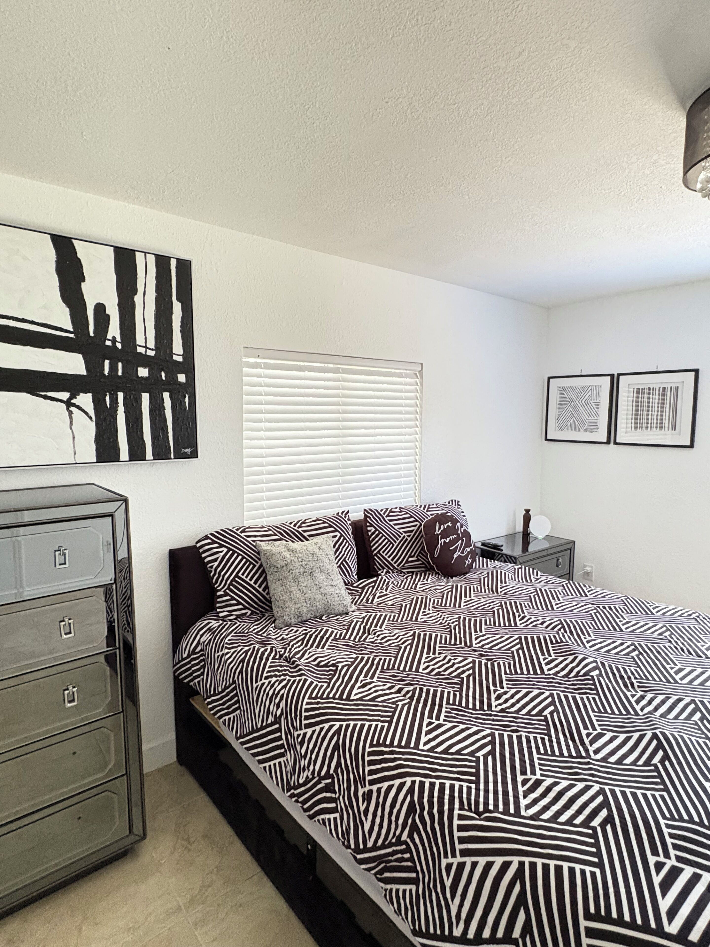A bedroom with a bed, a dresser, a window with blinds, and two framed pictures on the wall.