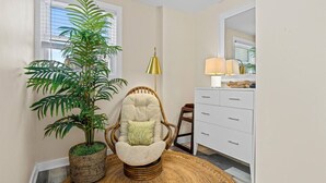 A room with a large potted plant, a wicker chair with a cushion, a white dresser, and a lamp.