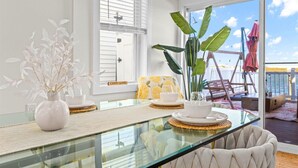 Un coin repas avec une table en verre, des chaises blanches, un coussin jaune et une grande plante.