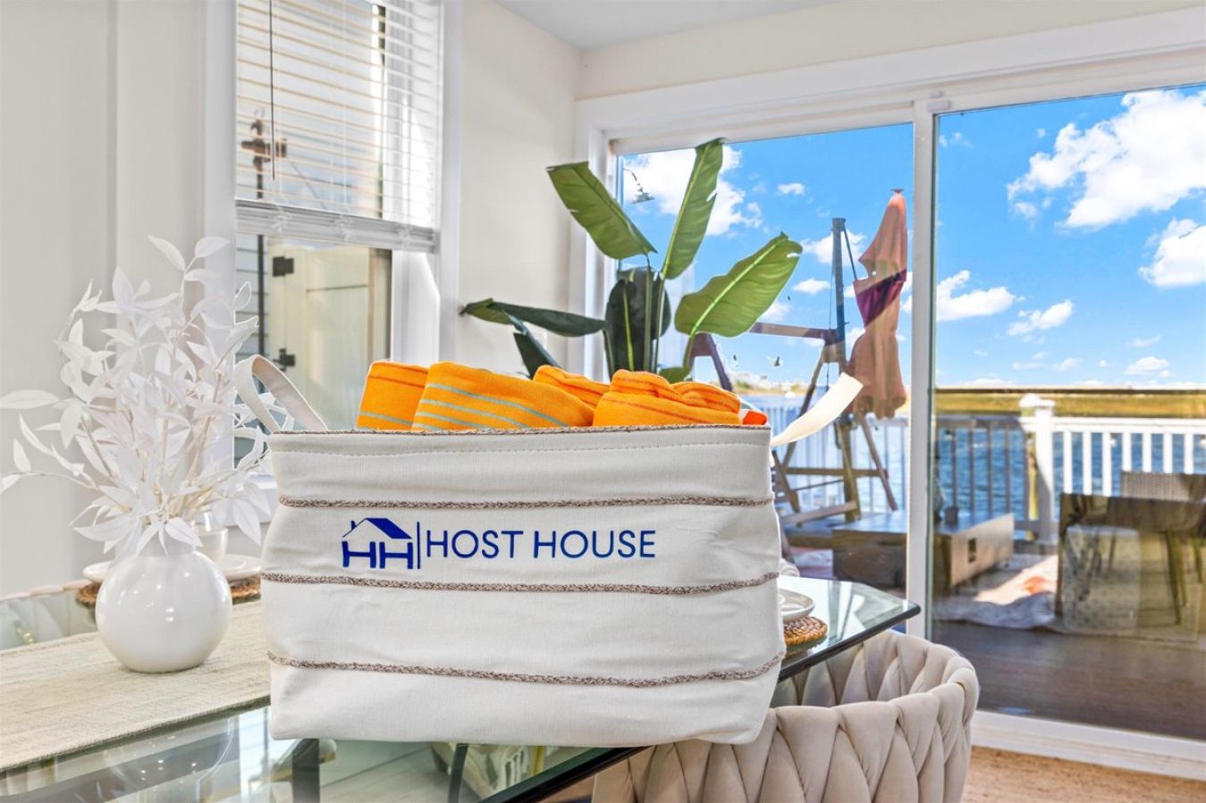 A white bag with "HOST HOUSE" branding on a glass table, a white vase with white flowers, and a view of a balcony with a chair and umbrella.