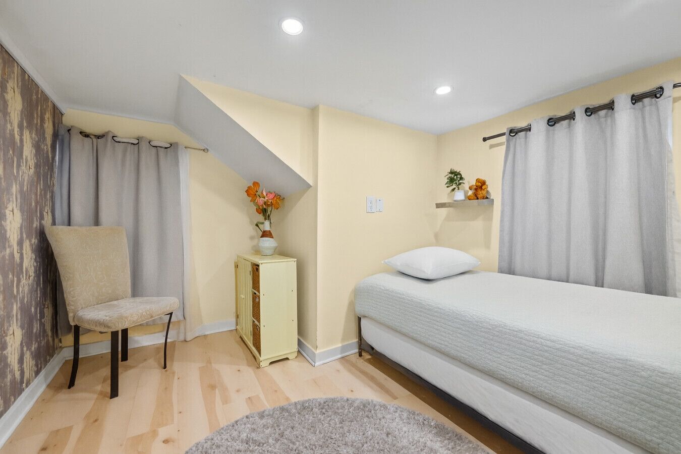 A bedroom with a bed, a chair, a small cabinet, and a shelf with a plant and a teddy bear.