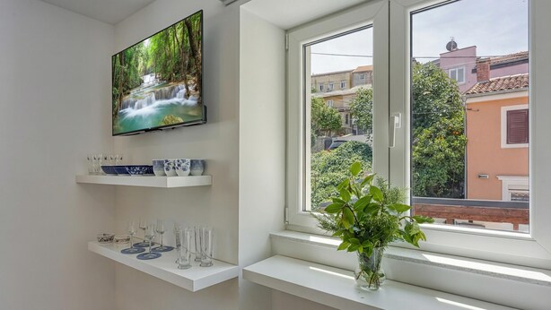 Une cuisine moderne avec un téléviseur fixé au mur, une fenêtre donnant sur le quartier et une étagère où sont disposés des verres et un vase avec des plantes.