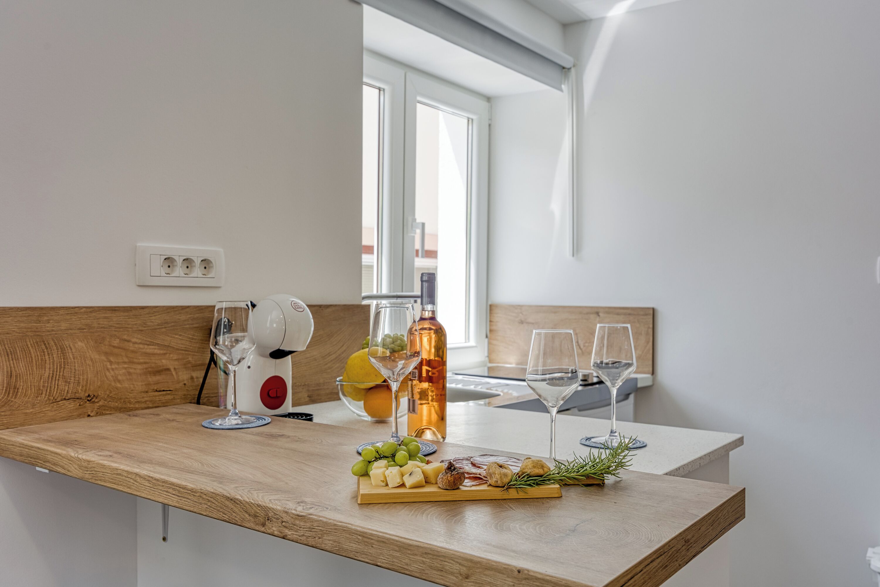 A kitchen counter with a wooden surface, a wine glass, a bottle of olive oil, and a cheese board with various items.