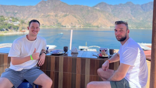 Two men sitting at a wooden table with a view of a lake and mountains in the background.