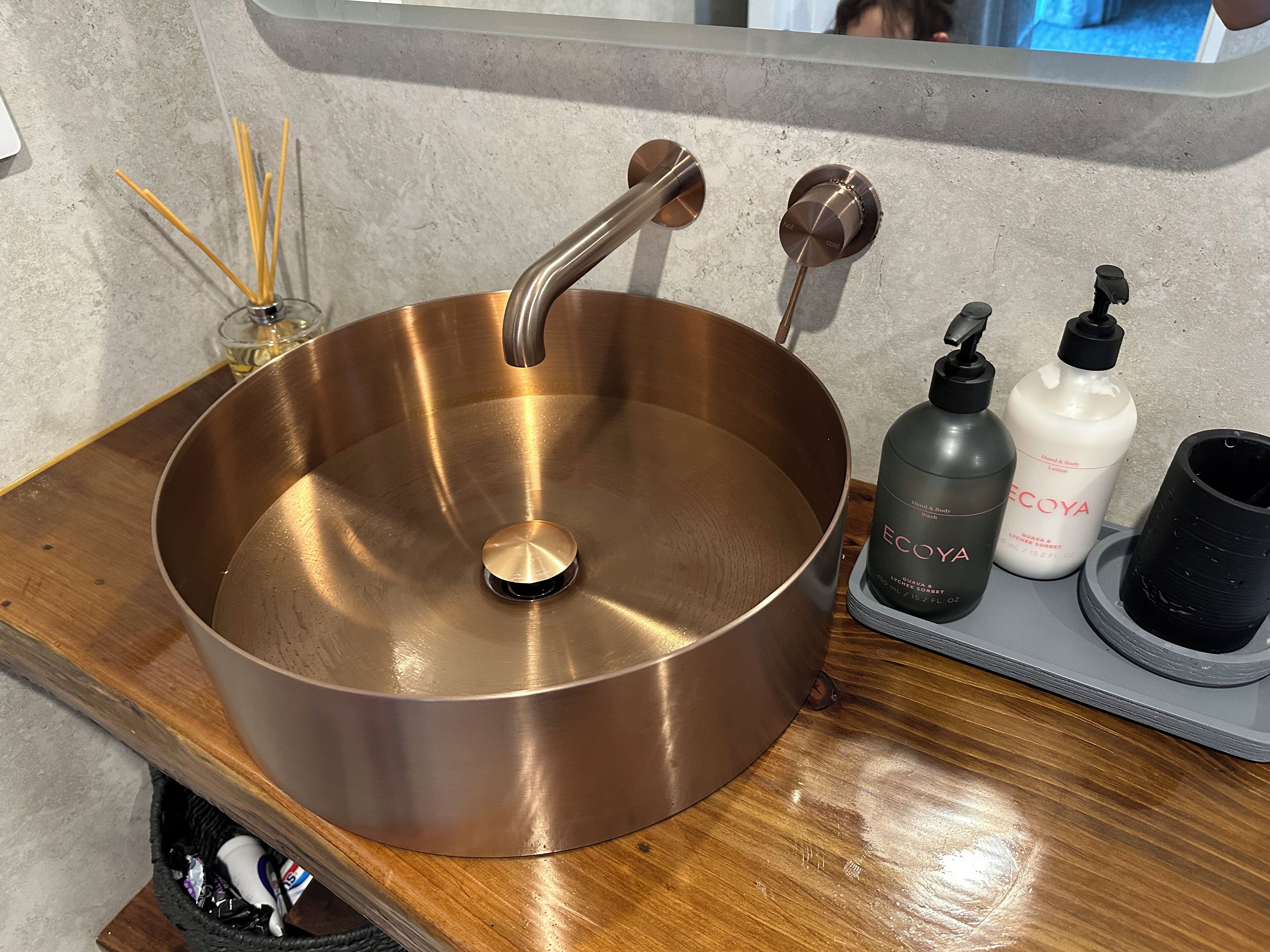 Ein Messingbecken mit modernem Wasserhahn, ein Glasbehälter mit Schilfdiffusor und zwei Flaschen Hand- und Körperlotion auf einer Holzauskleidung.