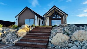 Une maison moderne avec une façade en pierre et de grandes fenêtres, un escalier en bois menant à l’entrée, et un ciel d’un bleu limpide.