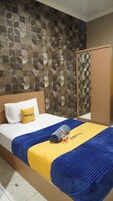 Una habitación de hotel con un papel tapiz estampado, una cama con cubrecama azul y amarillo, un armario de madera y un almohadón blanco con detalles amarillos.