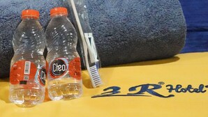 Una cama de hotel con una almohada amarilla y una manta con el logo del hotel. Dos botellas de agua y un cepillo de dientes están sobre la manta.