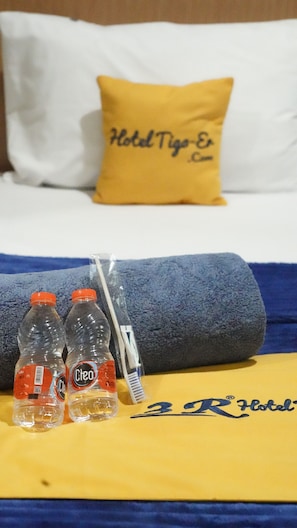 Una cama de hotel con una almohada amarilla y una manta con el logo del hotel. Dos botellas de agua y un cepillo de dientes están sobre la manta.