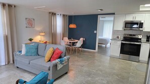 A modern living room with a grey sofa, colorful cushions, a dining table with chairs, a kitchen with a microwave and oven, and a blue accent wall. - Peaceful accomodation. Walking distant to Port Richey waterfront restaurants. (Port Richey)