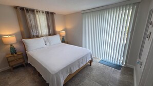 A bedroom with a bed, two bedside tables with lamps, a window with curtains, and a door with a handle. - Peaceful accomodation. Walking distant to Port Richey waterfront restaurants. (Port Richey)