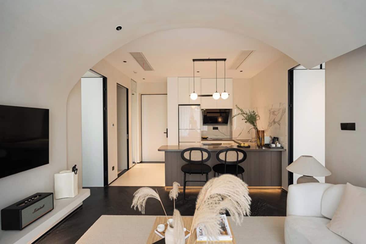 A modern living room with a flat-screen TV, a coffee table with decorative items, a sofa, and a dining area with chairs and a kitchen in the background.