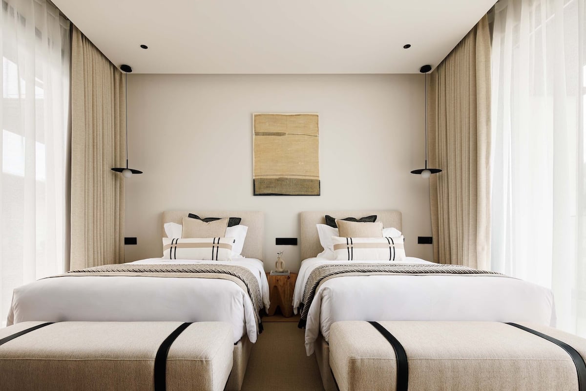 A hotel room with two beds, a large window with sheer curtains, a minimalist painting on the wall, and a small wooden nightstand between the beds.