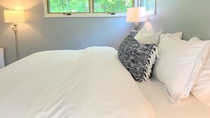 A bedroom with a large bed, two bedside lamps, a window with a view of trees, and a nightstand with a lamp.