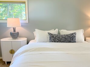 A neatly made bed with white bedding and a patterned pillow, a bedside lamp on a white nightstand, and a window with a view of trees.