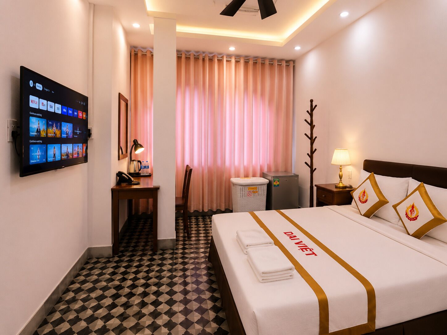 Une chambre d’hôtel comprenant un lit, un bureau, une télévision, une chaise, une poubelle et une table de chevet avec une lampe.