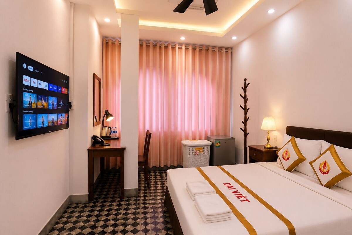 A hotel room with a bed, a desk, a television, a chair, a trash bin, and a nightstand with a lamp.