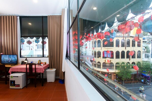 A hotel room with a large window, a table with a red and white checkered tablecloth, a chair, a small refrigerator, and a trash bin. - The Rice Hotel & Tour - 195 De Tham (Ho Chi Minh City)