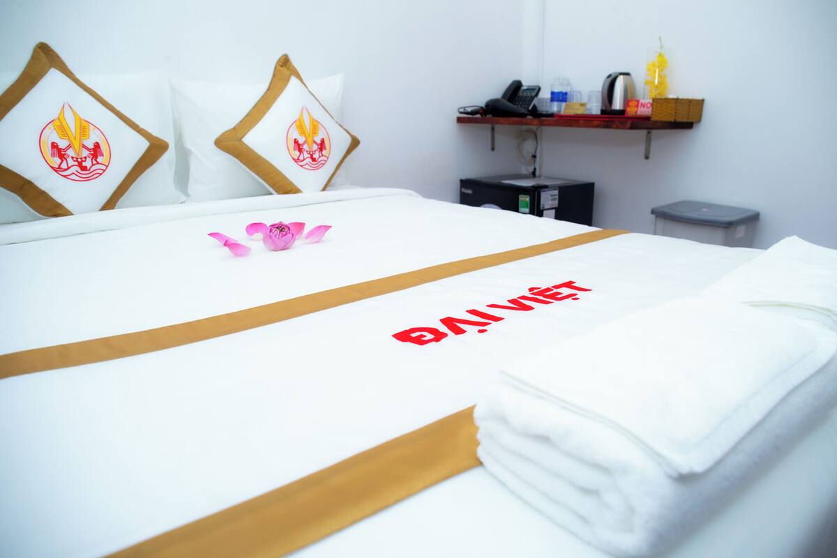A hotel room with a neatly made bed, pillows with a flame design, a small table with a phone and a basket, and a trash bin.