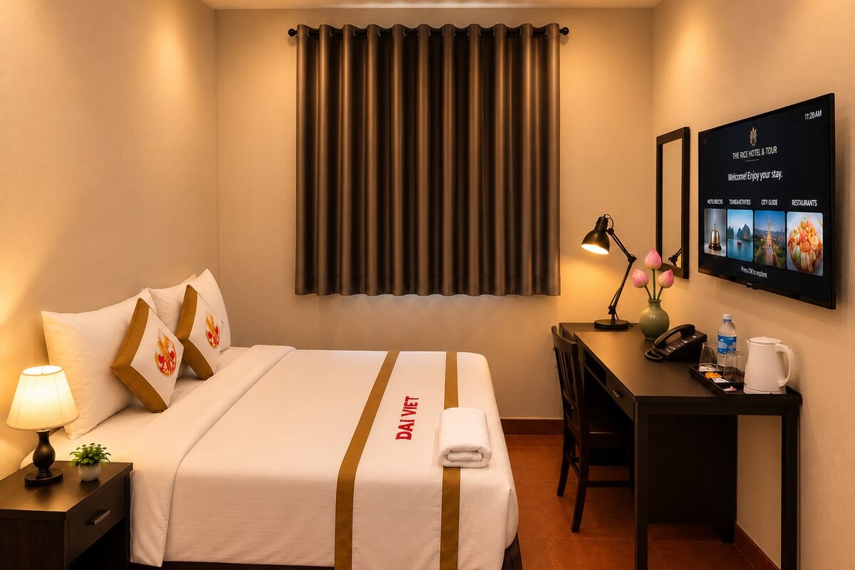 A hotel room with a bed, a desk, a chair, a nightstand, a lamp, a vase with flowers, a wall-mounted TV, and a curtain.
