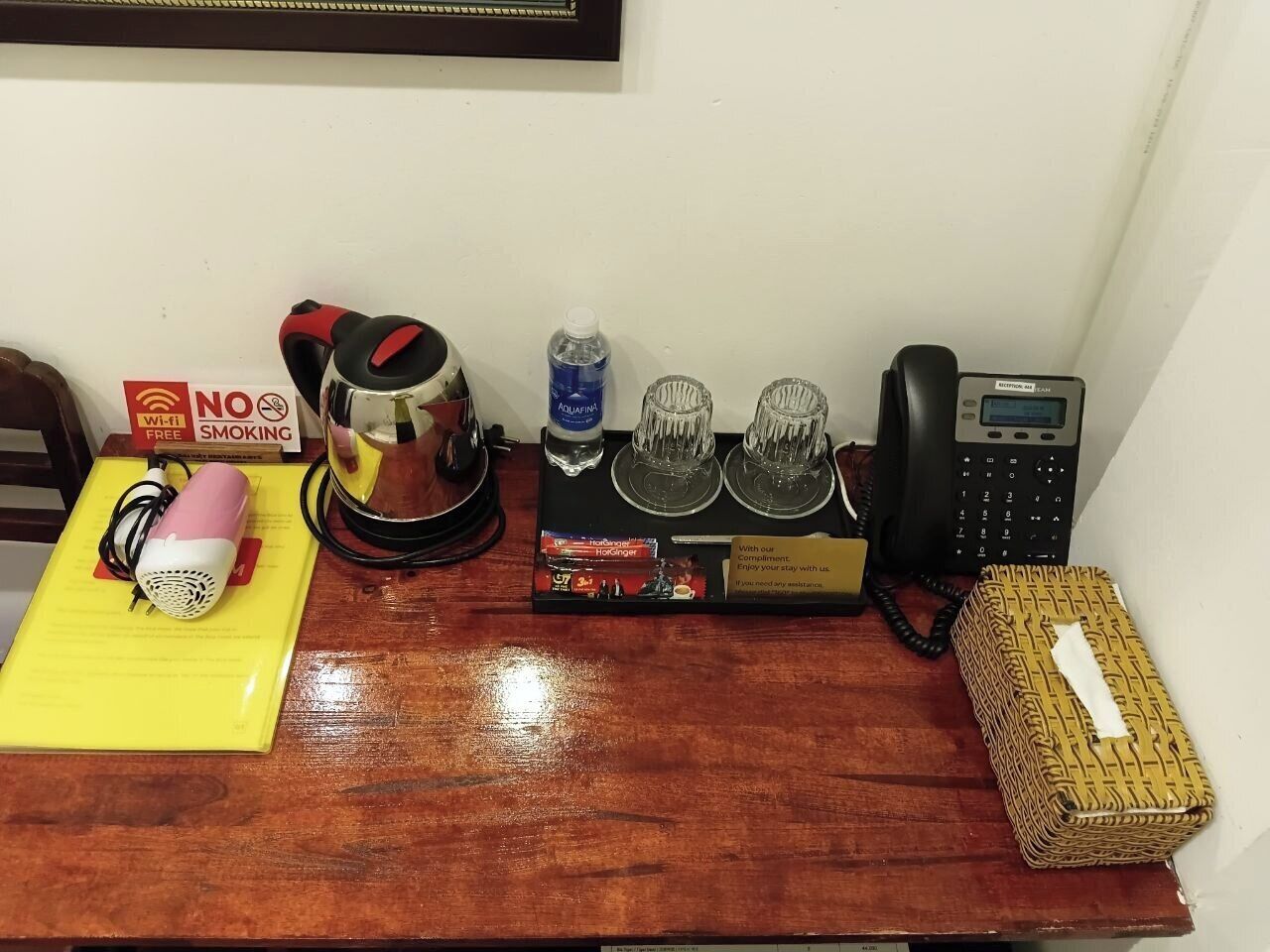 Un tavolo di legno con un asciugacapelli, un bollitore, una confezione di acqua Aquafina, due bicchieri, un telefono e un cesto decorativo.