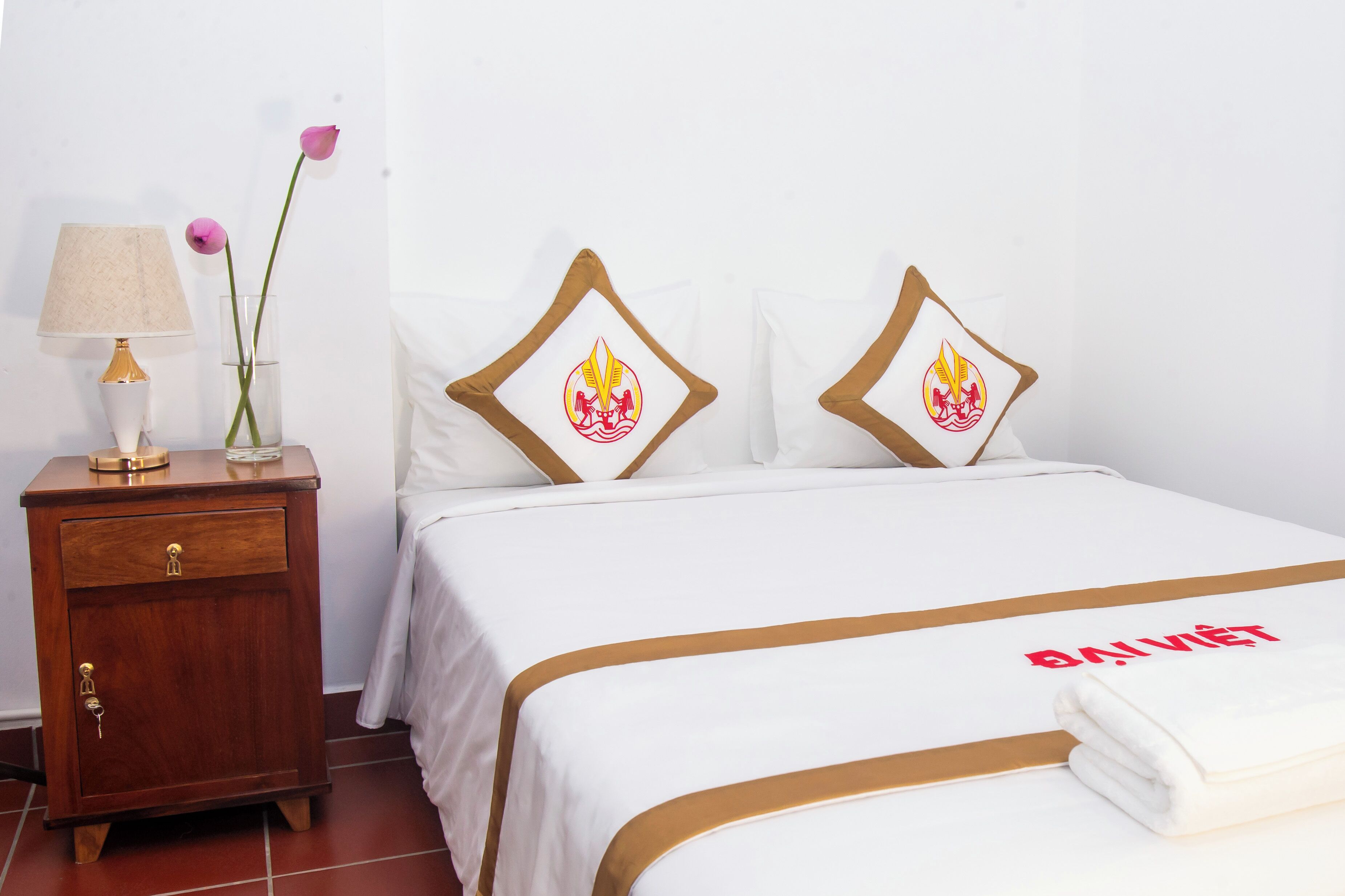 A neatly made bed with a white and gold bedspread, two pillows with a flame design, a bedside table with a lamp and a vase with flowers, and a folded towel on the bed.