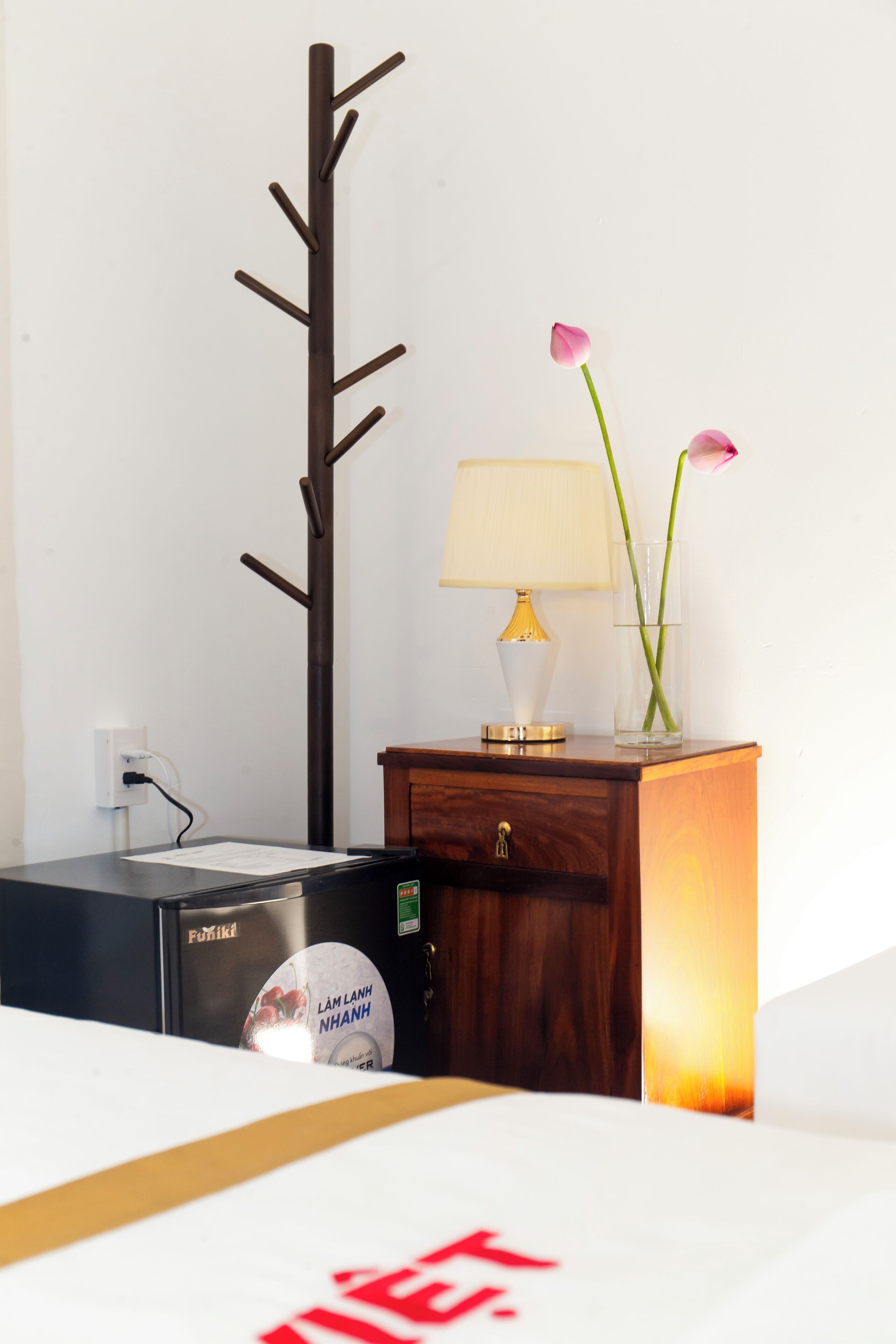 Een hotelkamer met een bed, een nachtkastje van hout met een lamp en bloemen, een kapstok en een minibar.