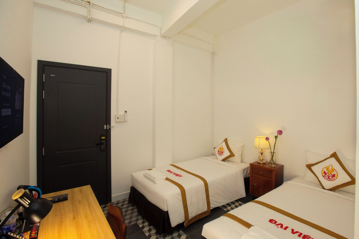 A hotel room with two beds, a desk with a lamp, a TV, and a door.
