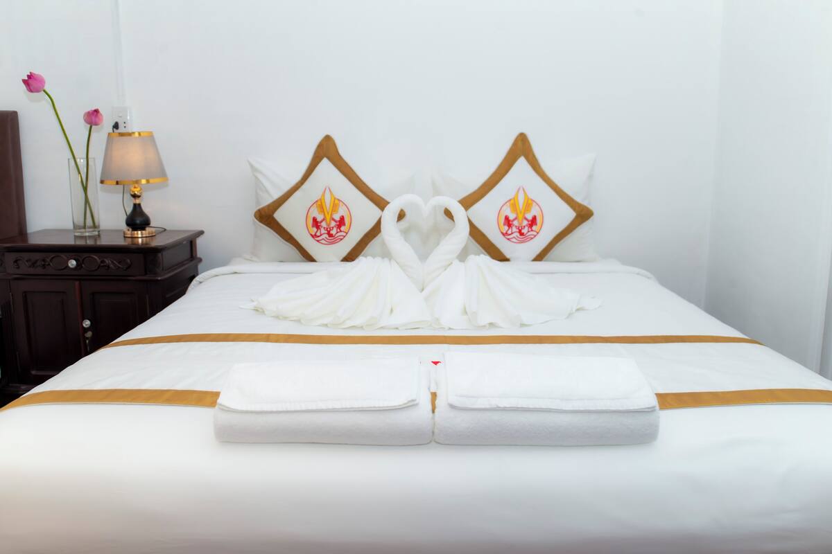 A neatly made bed with towels arranged in a heart shape, a bedside table with a lamp, and two pillows with a flame design.