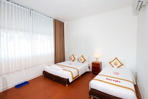 A hotel room with two beds, a window with curtains, a nightstand with a lamp, and a pair of slippers on the floor. - The Rice Hotel & Tour - 195 De Tham (Ho Chi Minh City)