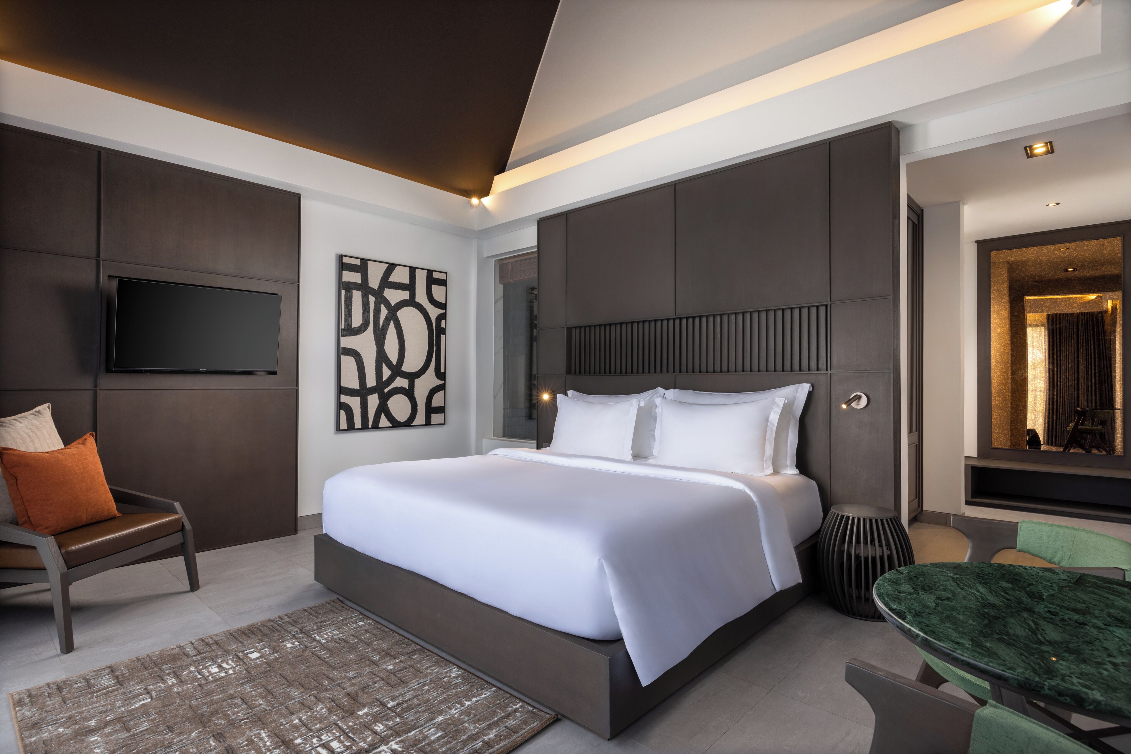 A modern hotel room with a large bed, a flat-screen TV mounted on the wall, a seating area with a chair and a table, and a decorative wall piece.